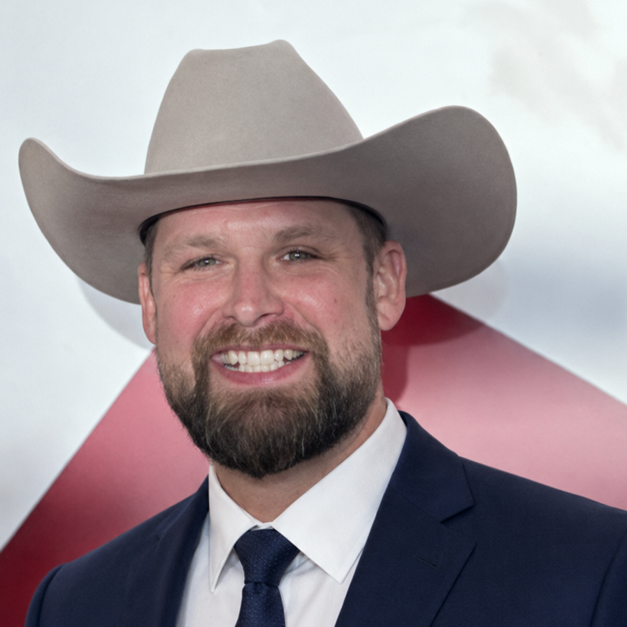 A smiling man wearing a cowboy hat standing for a picture in front of a red and white background.