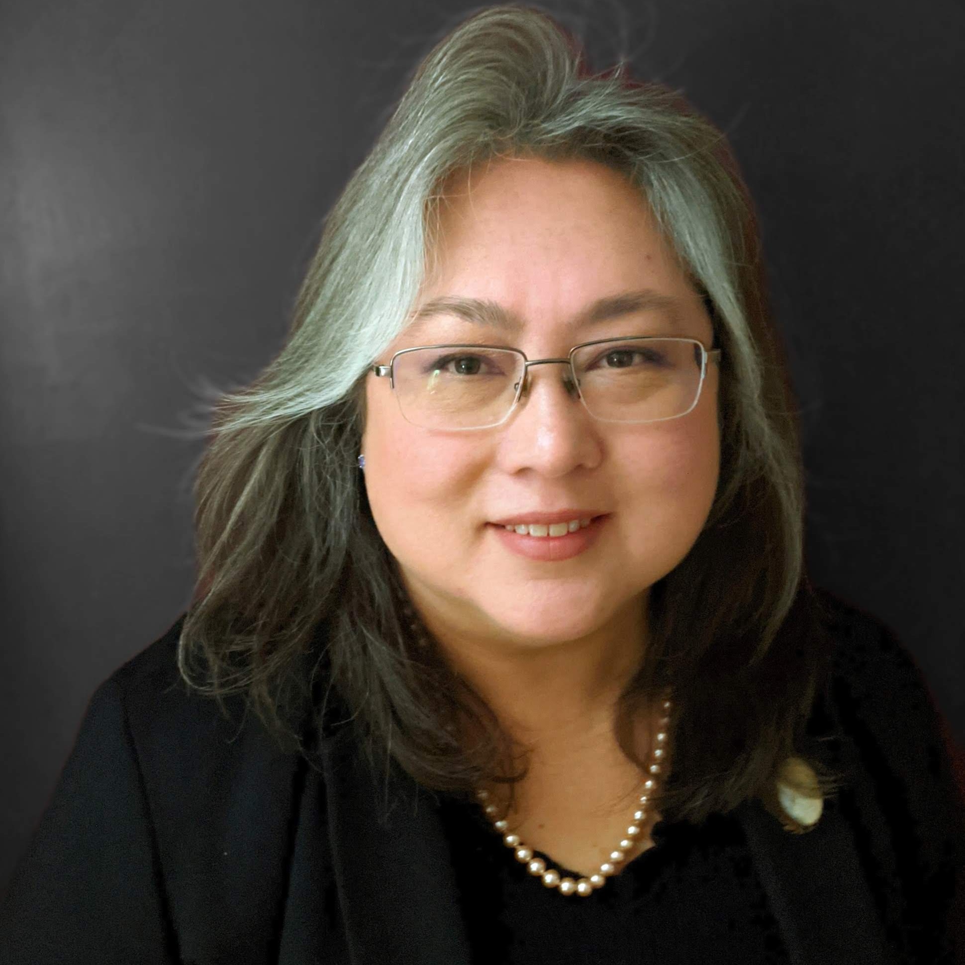 A smiling woman with long brown and gray hair standing in front of a dark wall.