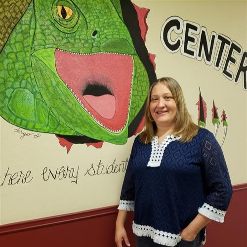 A smiling woman with shoulder length blonde hair in a blue and white shirt standing in front of a wall with an iguana painting.