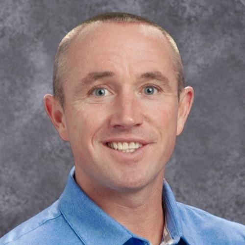 A smiling man with short hair in a blue shirt against a grey background.