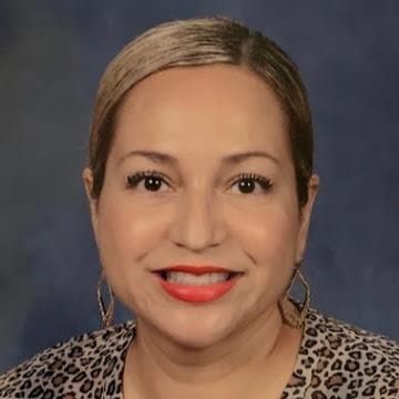 A smiling woman with brown hair and large earings wearing an animal print shirt against a gray background.