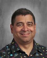 A smiling man with short dark hair wearing a colorful patterned shirt against a gray background.