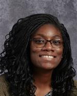 A smiling woman with long, curly hair wearing glasses and a sweater, set against a gray background.