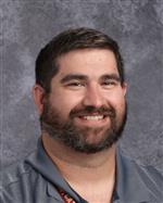 A smiling man with a beard, wearing a gray polo shirt, poses for a professional headshot against a textured background.