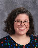A smiling woman with curly hair and glasses, wearing a colorful patterned shirt, poses for a portrait against a gray background.