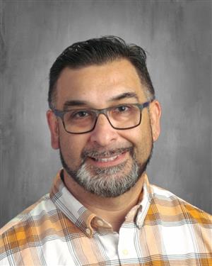 A smiling man with a beard and glasses, wearing a plaid shirt, poses for a professional headshot against a gray background.