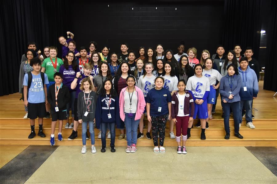 A large group of students, approximately 50 in total, stands together on a stage with wooden flooring, smiling and posing for a photo, while wearing various casual outfits and name tags.