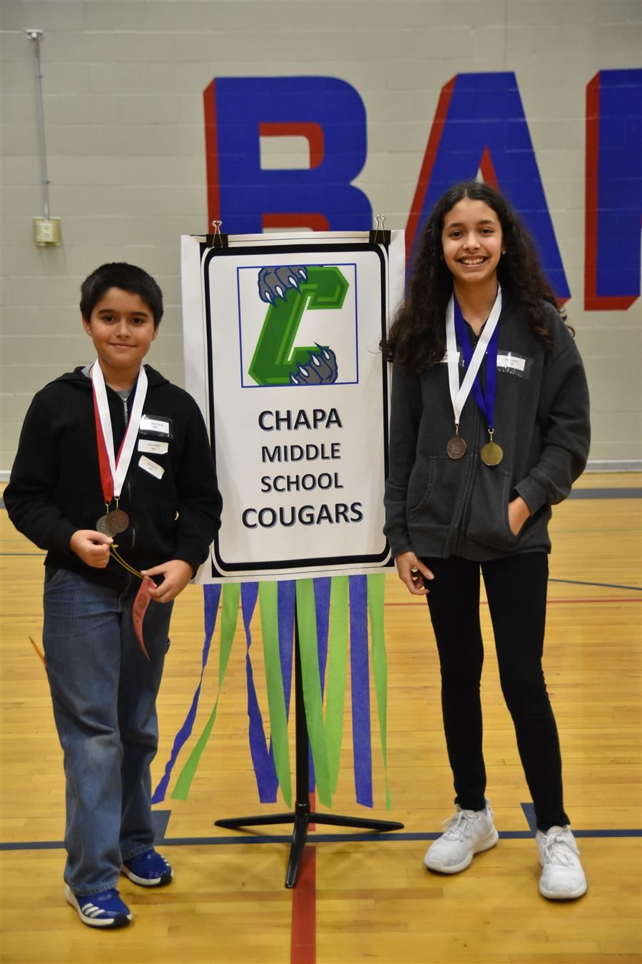 Two students stand beside a sign that reads 'Chapa Middle School Cougars,' each wearing medals and smiling in a gymnasium setting.