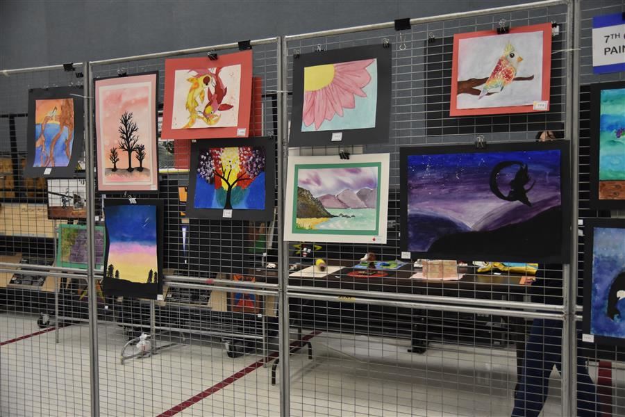 A display of various framed artworks hung on wire mesh panels in an exhibition space, showcasing colorful paintings and drawings.