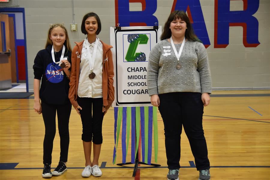Three middle school students stand together, each wearing a medal, in front of a sign that reads 'Chapa Middle School Cougars.'