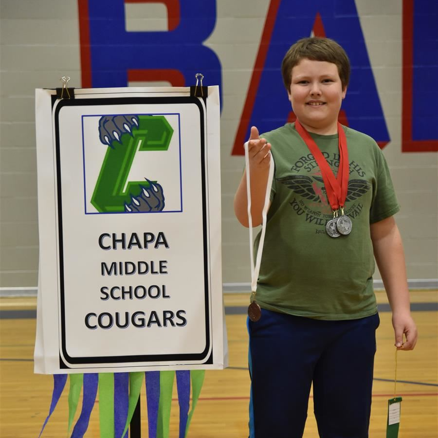 A young boy stands proudly next to a sign that reads 'Chapa Middle School Cougars,' wearing medals around his neck and holding a ribbon in his hand, in a gymnasium setting.