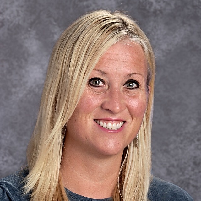 A smiling woman with long blonde hair, wearing a gray top, poses for a professional photo against a textured gray background.