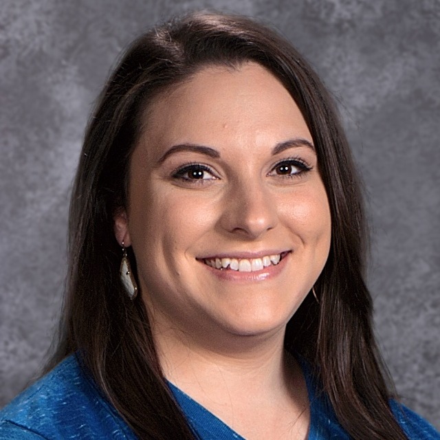 A smiling woman with long dark hair, wearing a blue shirt, is posed against a gray background.