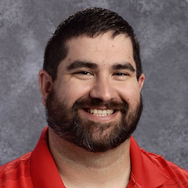 A smiling man with a beard, wearing a gray polo shirt, poses for a professional headshot against a textured background.