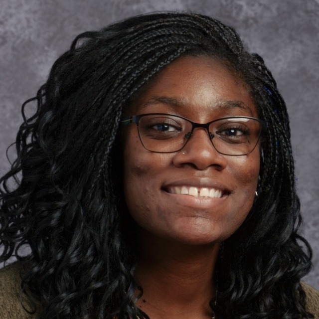 A smiling woman with long, curly hair wearing glasses and a sweater, set against a gray background.