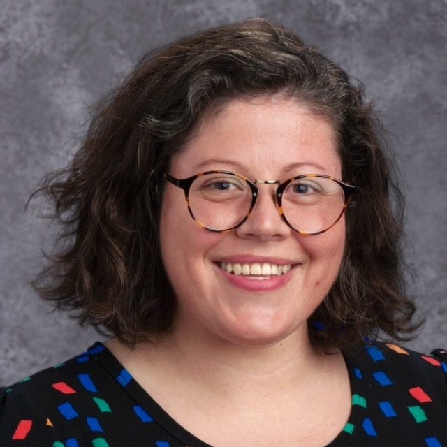 A smiling woman with curly hair and glasses, wearing a colorful patterned shirt, poses for a portrait against a gray background.