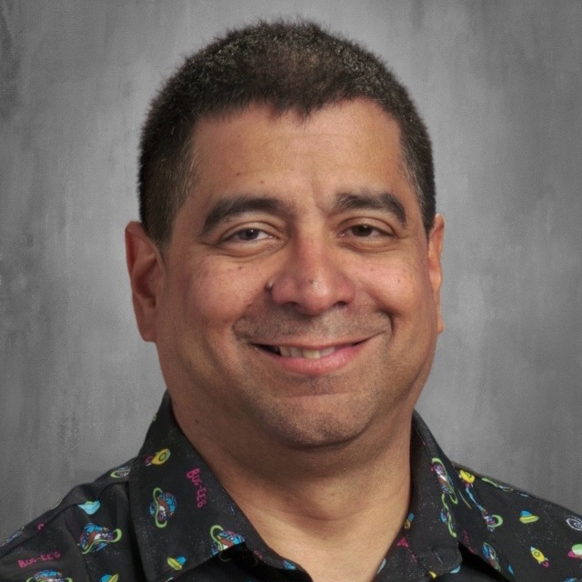 A smiling man with short dark hair wearing a colorful patterned shirt against a gray background.