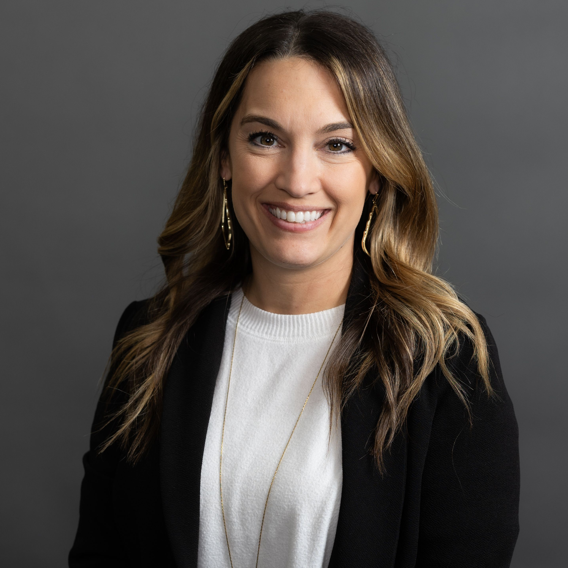 A smiling woman with long brown hair with blonde highlights, wearing a black jacket and a white top while standing in front of a professional dark grey photography backdrop.
