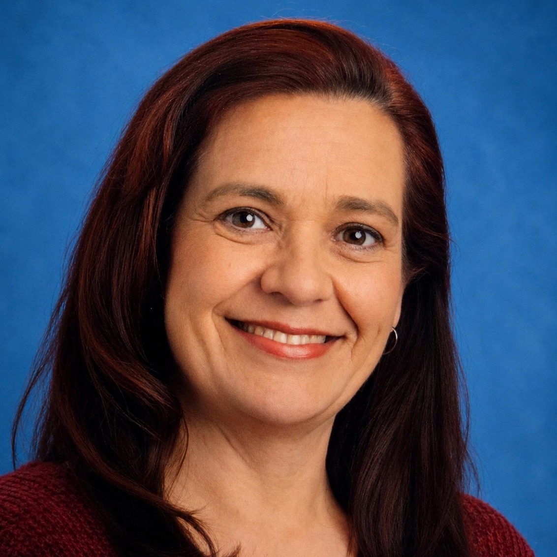 A smiling woman with dark brown hair in a red sweater posing for a professional photo taken in front of a blue background.