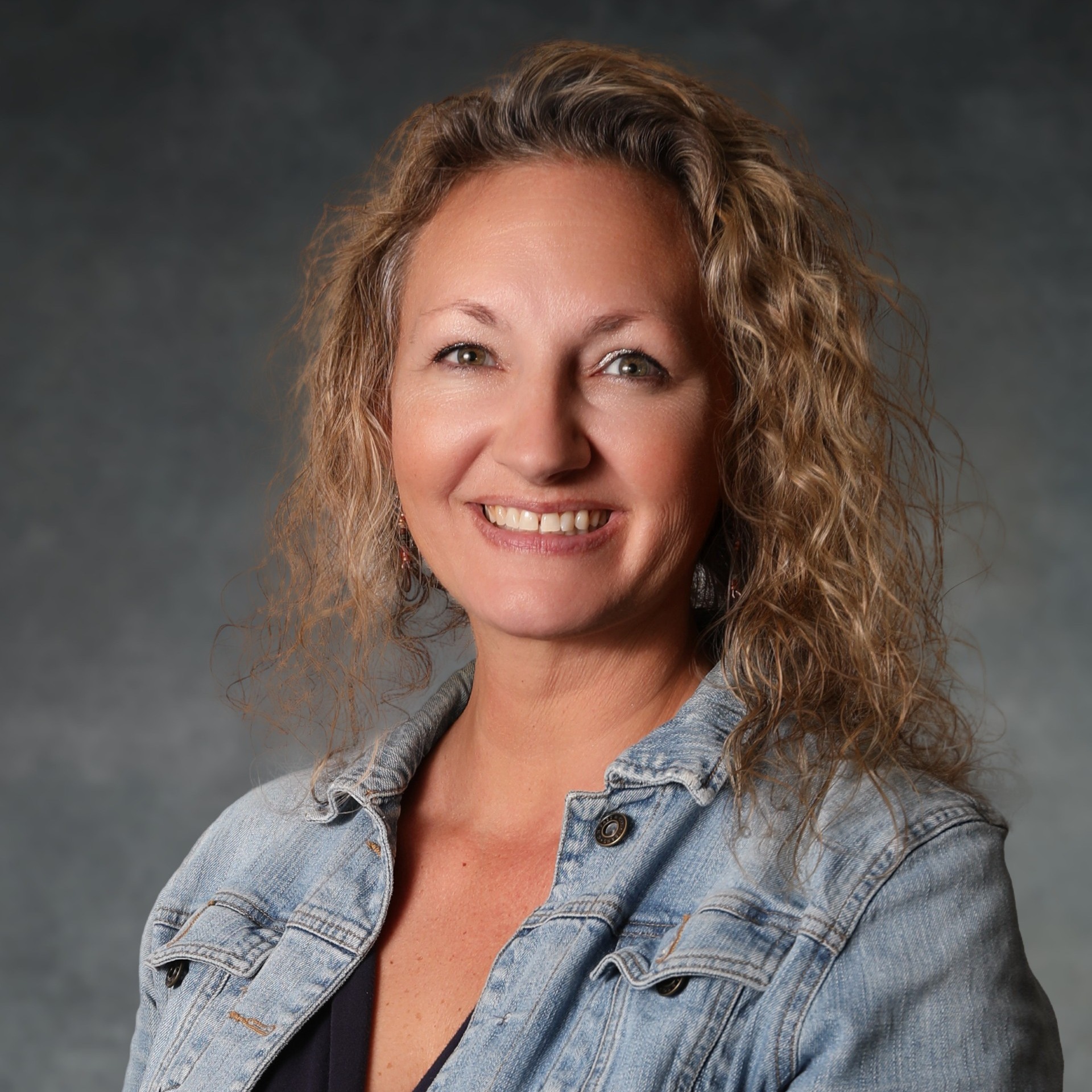 A smiling woman with shoulder length, curly, blonde hair in a denim jacket while in front of a dark grey photography background.