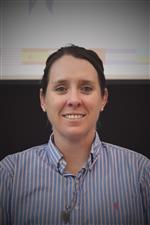 A woman with dark hair, wearing a blue and white striped shirt, smiles at the camera against a dark background.