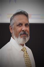 A man with a gray beard and mustache, wearing a white shirt and a yellow patterned tie, poses for the camera against a blurred background.