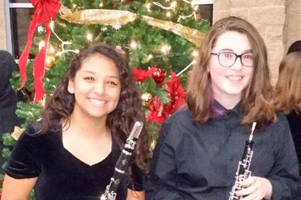 Two young musicians stand in front of a decorated Christmas tree, each holding a woodwind instrument, smiling for the camera.