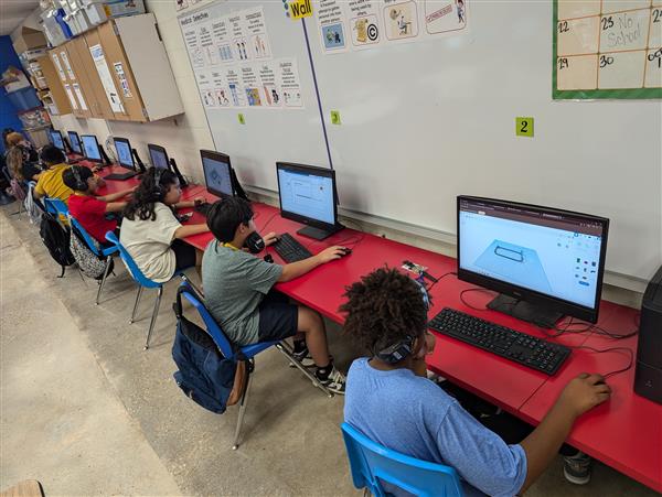 A classroom filled with students working on computers at red tables, focused on their screens, with educational materials visible on the walls.