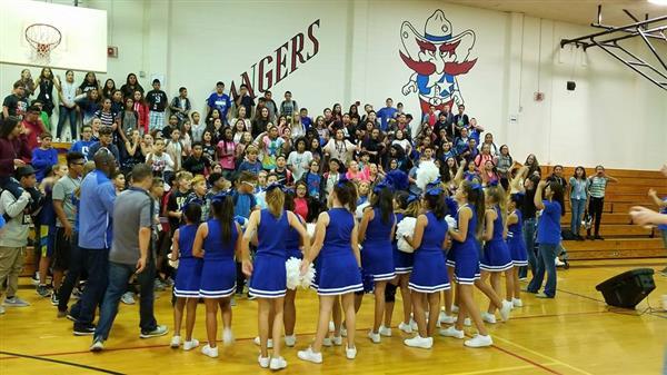 A group of cheerleaders in blue uniforms gathers in a gymnasium surrounded by an audience, with a large mural of a mascot on the wall behind them.