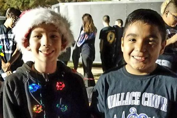 Two children are smiling at the camera, one wearing a Santa hat and a shirt with colorful lights, while the other is dressed in a dark shirt with a logo and has a red sweater tied around his waist.