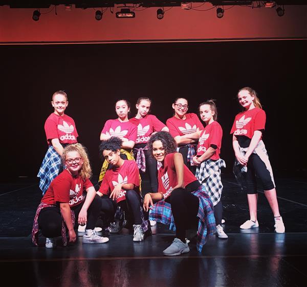A group of nine young dancers poses on stage, wearing matching red shirts and black pants, with some sporting plaid shirts tied around their waists, against a dark background.