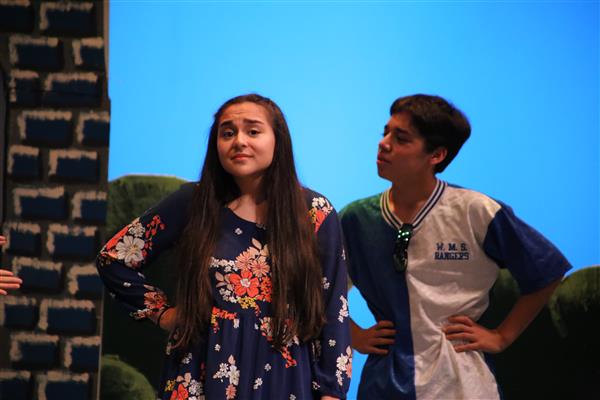 Two young actors perform on stage, with one wearing a floral dress and the other in a sports jersey, set against a bright blue background.