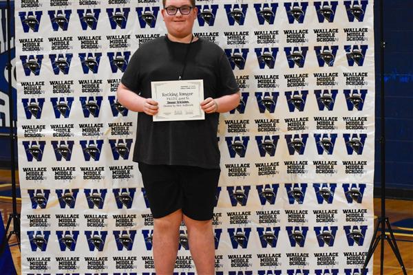 A young person stands proudly holding a certificate in front of a backdrop featuring repeated logos, wearing a black outfit and glasses.