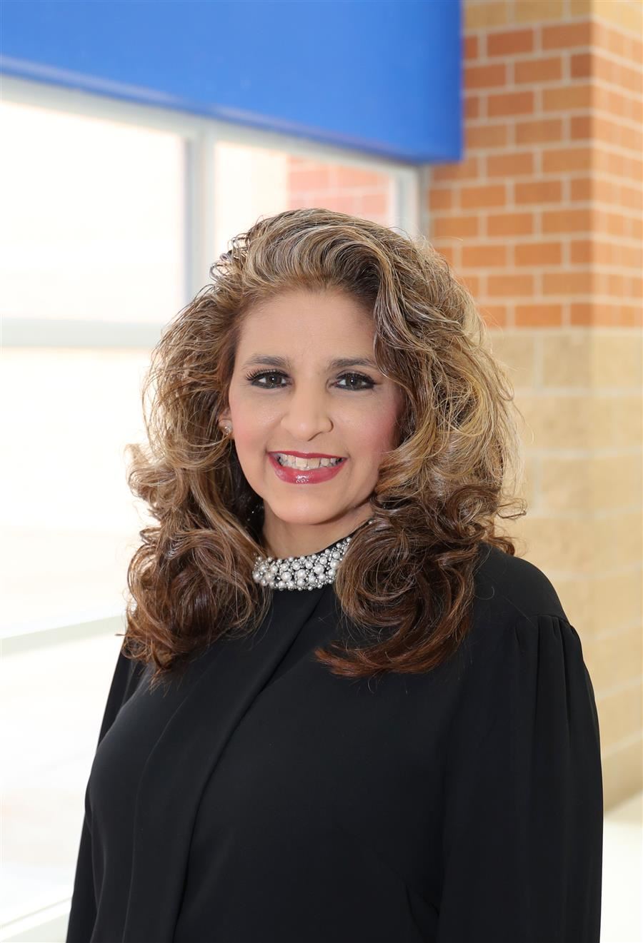 A woman with long, curly hair wearing a black outfit and a pearl necklace smiles in front of a window with a blue and brick background.