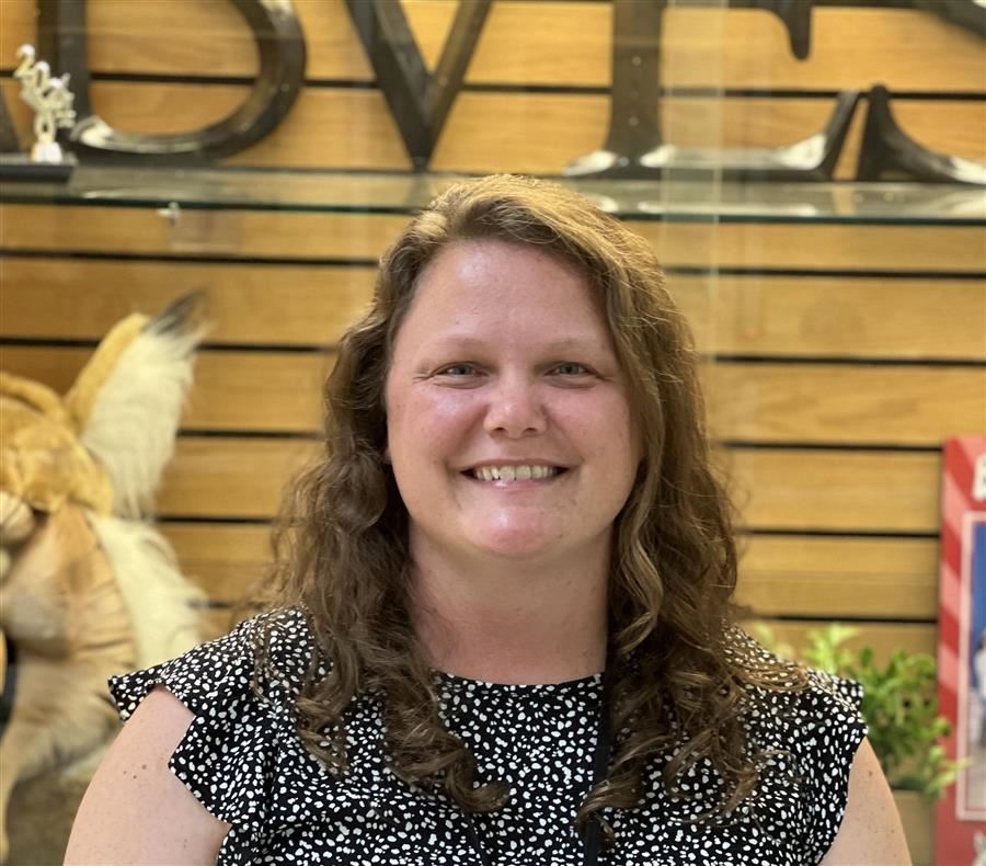 A smiling woman with curly hair is standing in front of a wooden wall with the word 'DVES' displayed, alongside a taxidermy animal in the background.