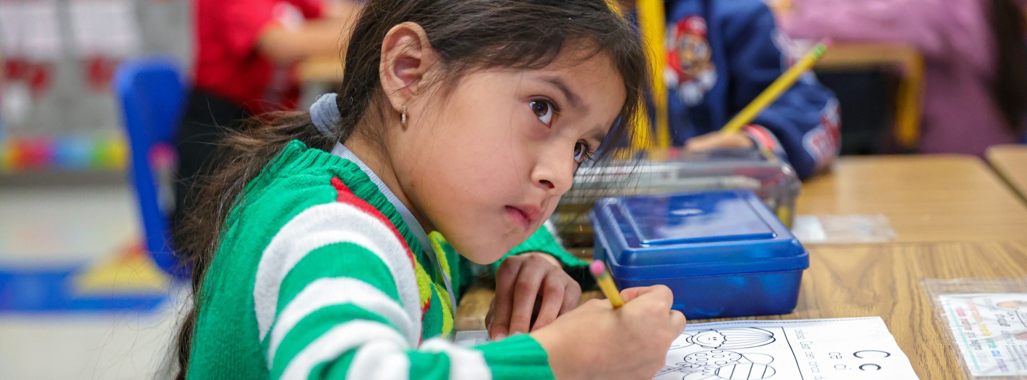 A young girl in a green and white sweater writing with a pencil and looking off camera.