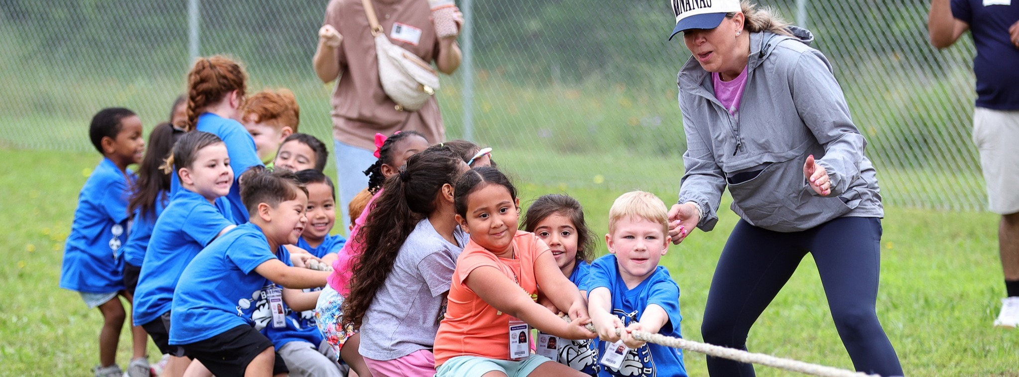 A large group of kids playing tug of war while adults cheer them on.