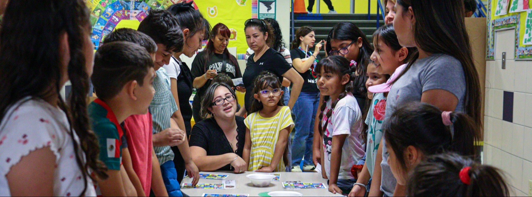 A group of Camino Real Elementary students and parents standing around a rectangular table with loteria cards on top. 