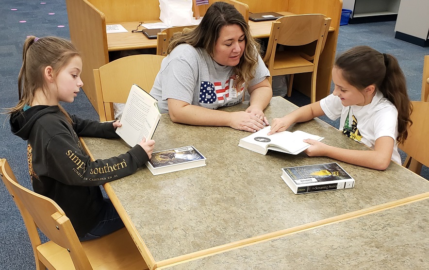 Three young girls engrossed in their reading with a woman standing by to assist them.