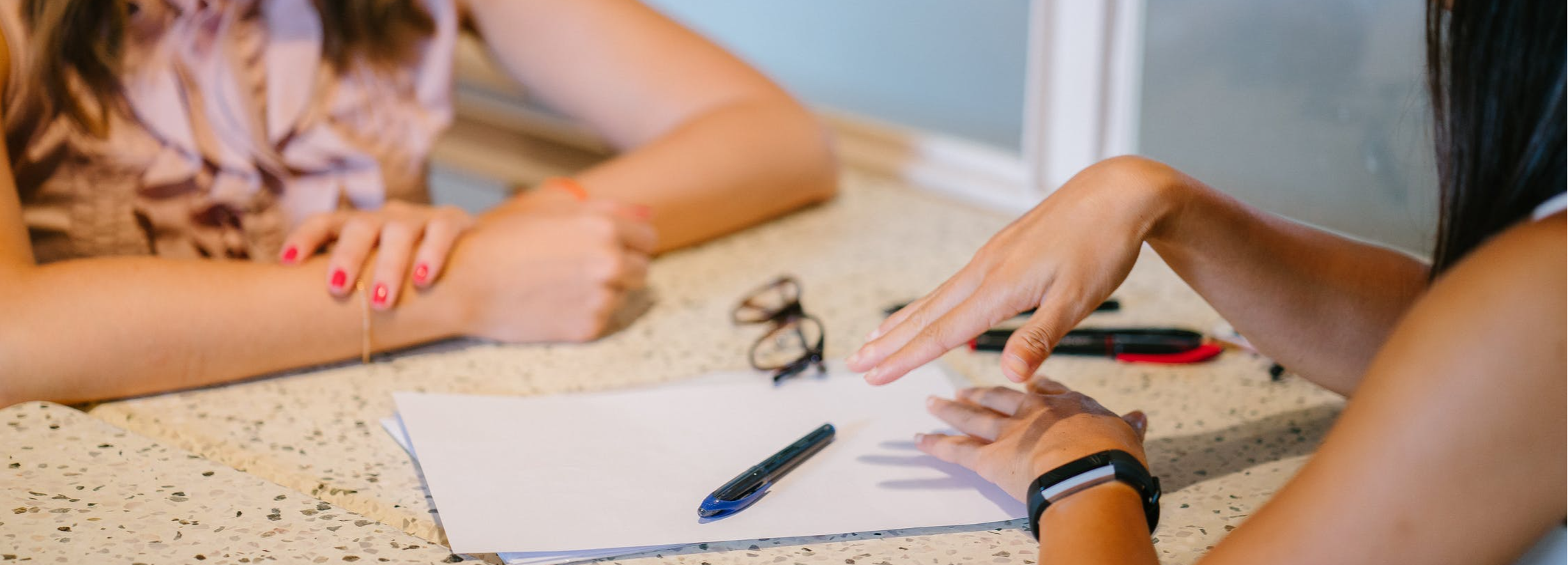 Two women engaged in conversation with papers and pen on the table.