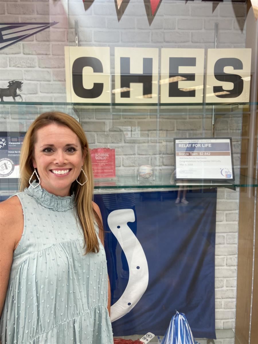 A woman with long hair wearing a light blue dress stands next to a display case featuring the letters "CHES" and various memorabilia, including a blue flag and a horseshoe.