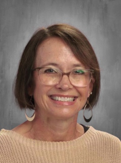 A smiling woman with short brown hair and glasses, wearing a beige sweater and large earrings, poses against a gray background.