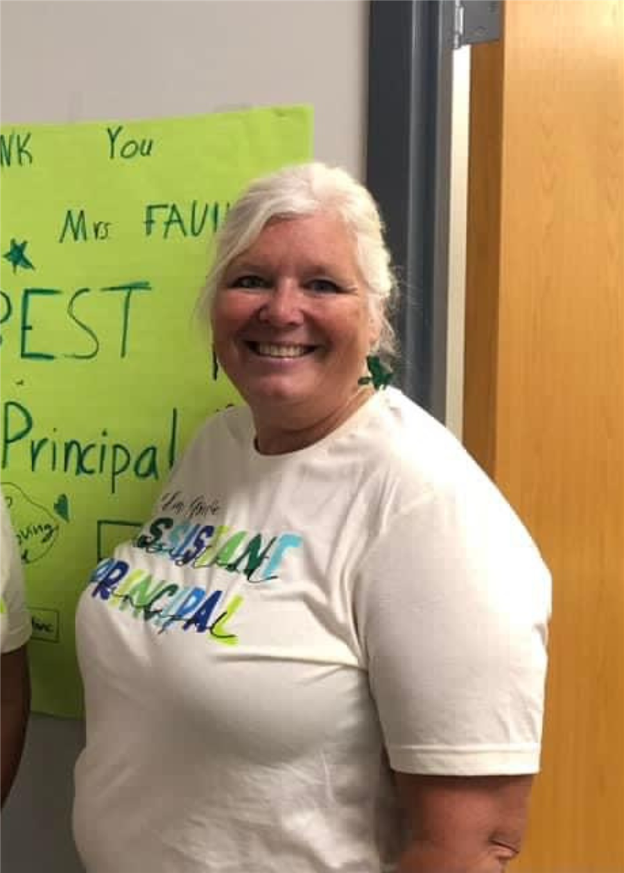 A smiling woman with short, light-colored hair stands in front of a colorful poster that expresses gratitude, wearing a white t-shirt with the words 'Best Principal' printed on it.