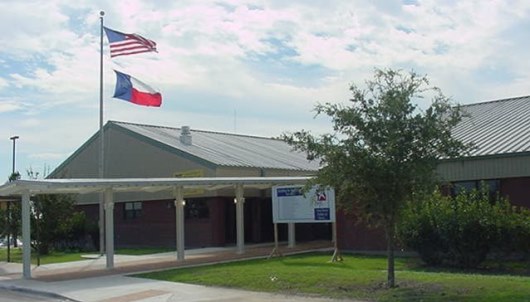 A building with a metal roof, featuring the American and Texas flags flying above it, surrounded by grass and trees under a cloudy sky.