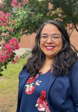 A smiling woman with long, wavy black hair and glasses stands outdoors in front of flowering plants, wearing a navy blue blouse with floral patterns.