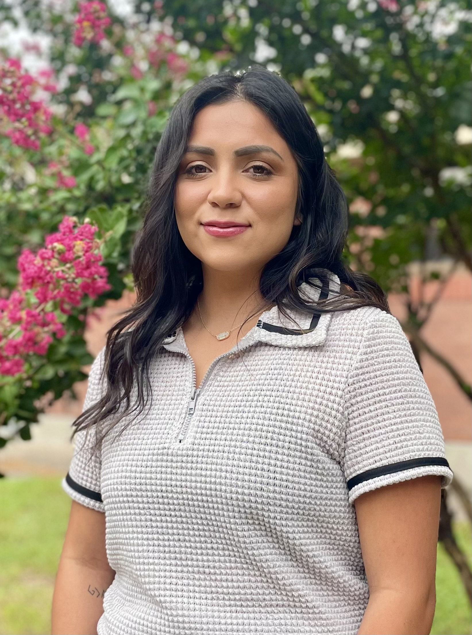 A woman with long dark hair stands outdoors in front of flowering plants, wearing a light-colored polo shirt with a zipper.