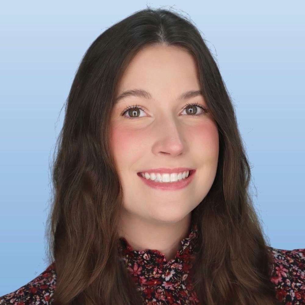 A woman with long brown hair smiling while in front of a light blue professional background.
