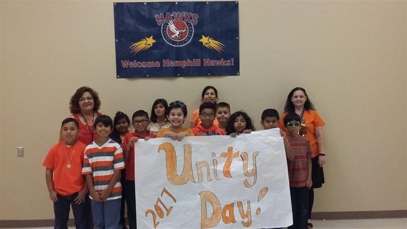 A group of children and adults, wearing orange shirts, stand together holding a large banner that reads 'Unity Day 2017' in a school setting with a welcoming banner in the background.
