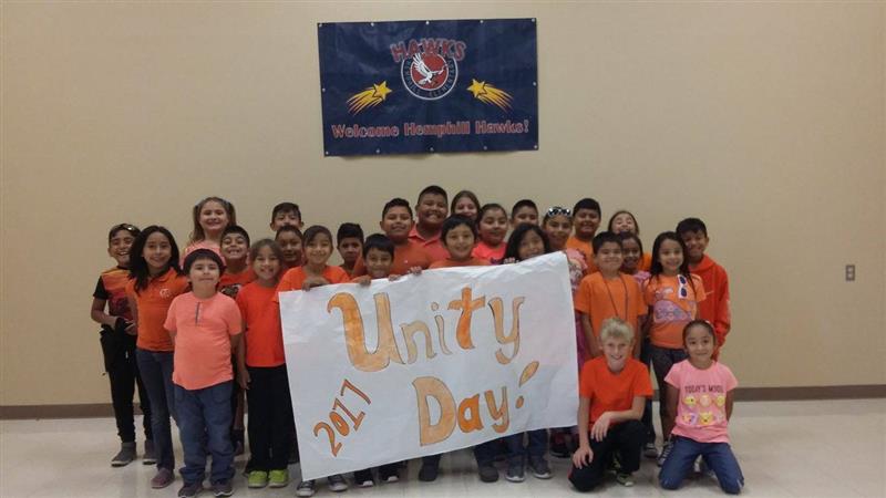 A group of children wearing orange shirts stands together in front of a banner, holding a sign that reads 'Unity Day 2017.'