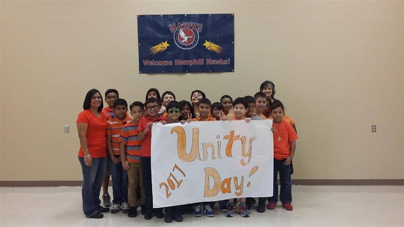A group of students and adults, all wearing orange shirts, stand together holding a large sign that reads 'Unity Day 2017' in a school setting, with a banner in the background.